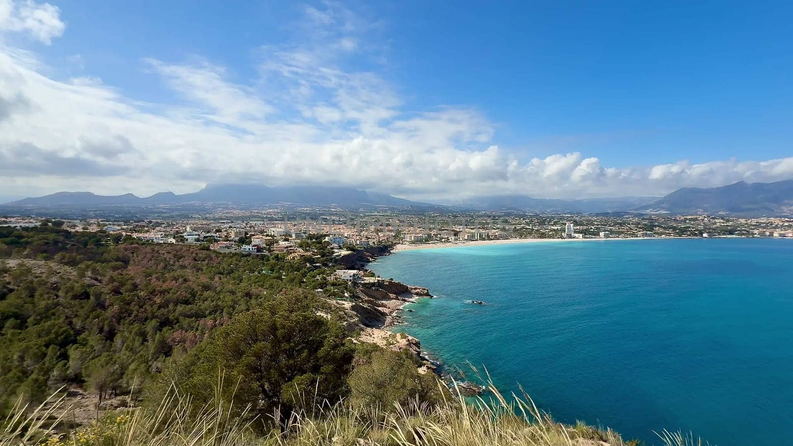 A stunning coastal landscape shows clear blue waters alongside majestic mountains under a bright, sunny sky