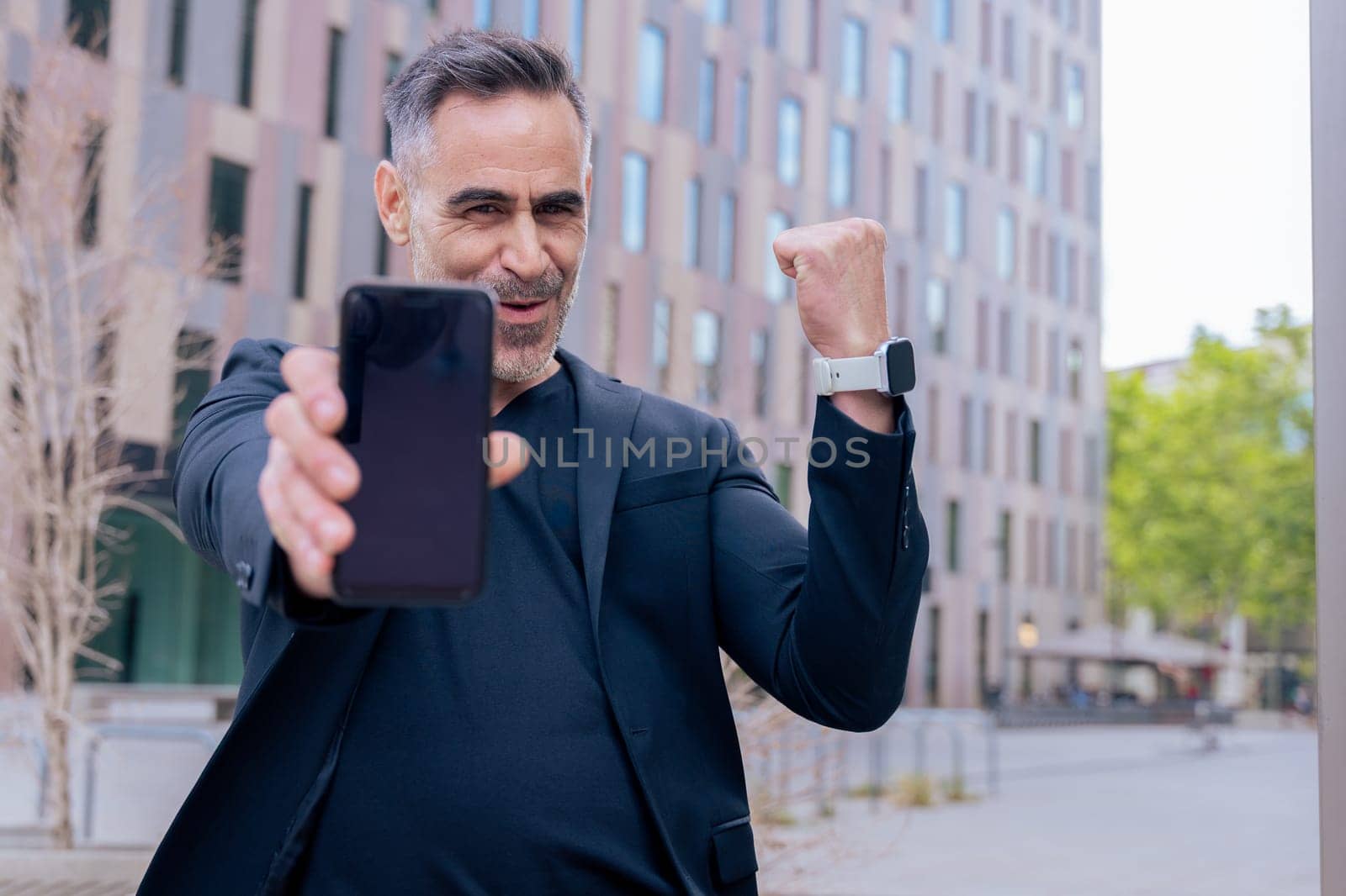 Happy man in black celebrates winning an online bet, holding up his smartphone and raising his fist in victory. He stands in a modern city area, full of joy and excitement.