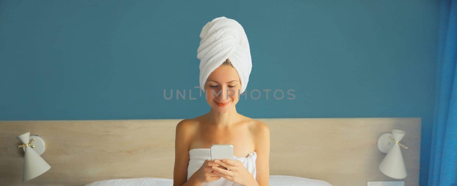 Happy young woman looking at mobile phone drying her hair after bath with towel on her head sitting on white bed in bedroom in morning