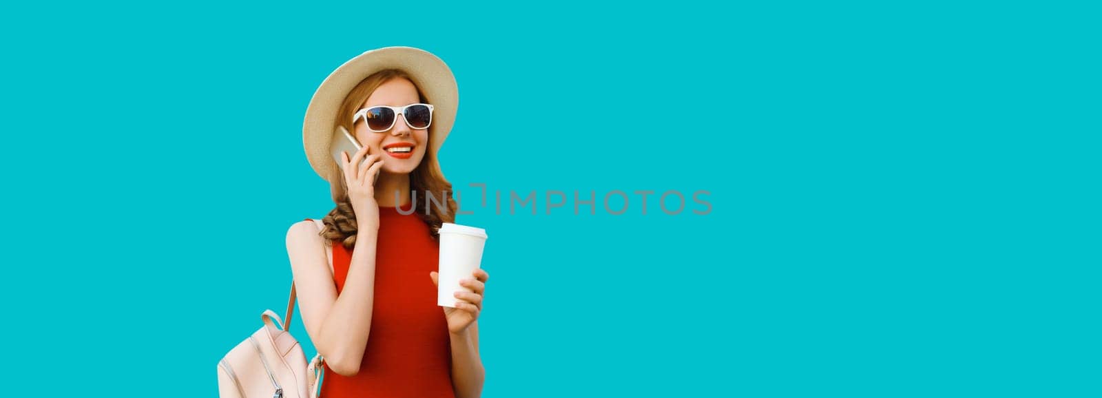 Portrait of happy cheerful young woman calling on phone with cup of coffee looking away in tourist hat with backpack on blue background
