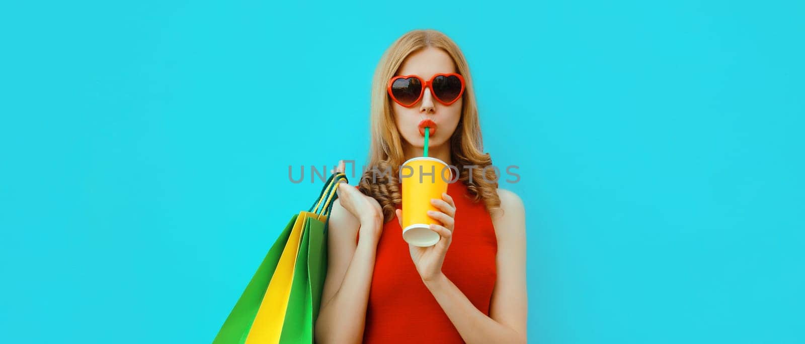 Portrait of beautiful relaxed young woman model with colorful shopping bags drinking juice in red heart shaped glasses on blue studio background