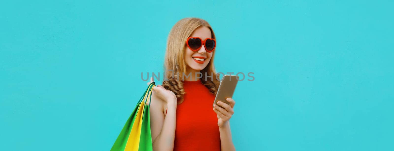 Beautiful happy young woman looking at smartphone with shopping bag in red dress with heart shaped sunglasses, girl holding phone on blue studio background