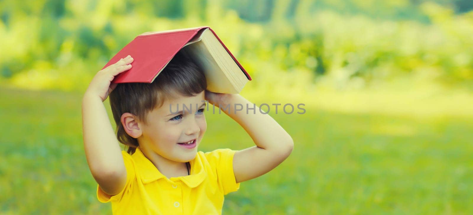 Happy little boy child reading a book sitting on green grass in summer park