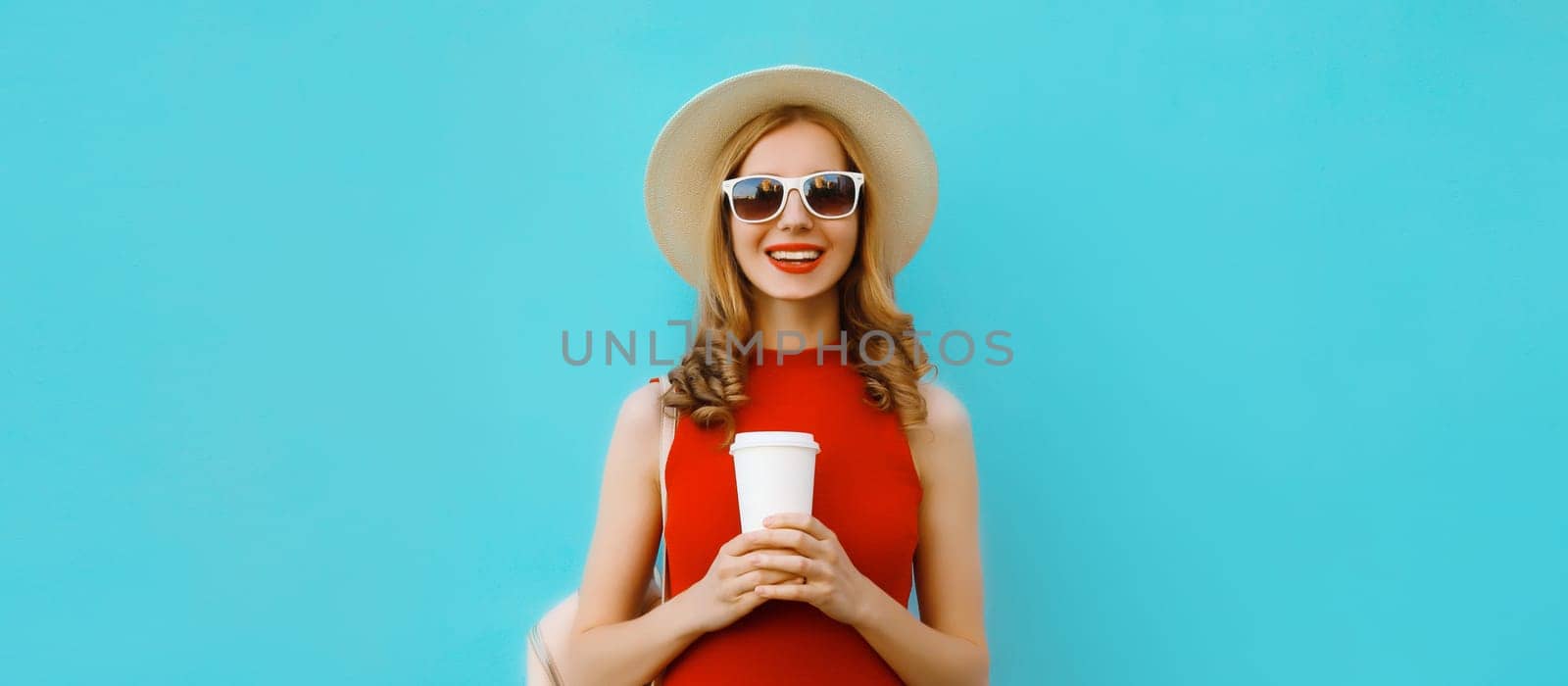 Portrait of beautiful happy young woman holding cup of coffee in summer tourist straw hat, red dress and glasses, pretty girl smiles with backpack on bright blue background