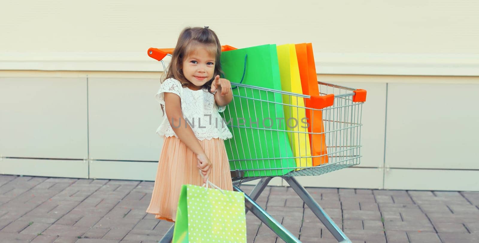 Happy little girl child with trolley cart and colorful shopping bags posing on city street