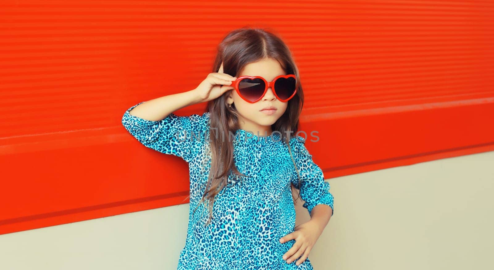 Portrait of beautiful little girl child in red heart shaped sunglasses and blue dress on city street on red background