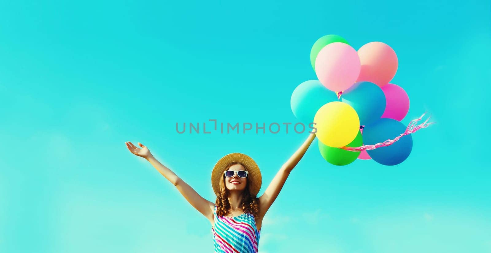 Happy joyful young woman with bunch of colorful balloons in summer straw hat having fun on sunny blue sky background
