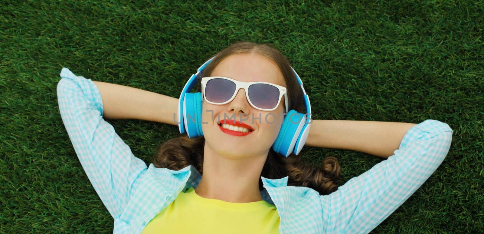 Portrait of happy smiling young woman listening to music in headphones while lying on green grass lawn in summer park