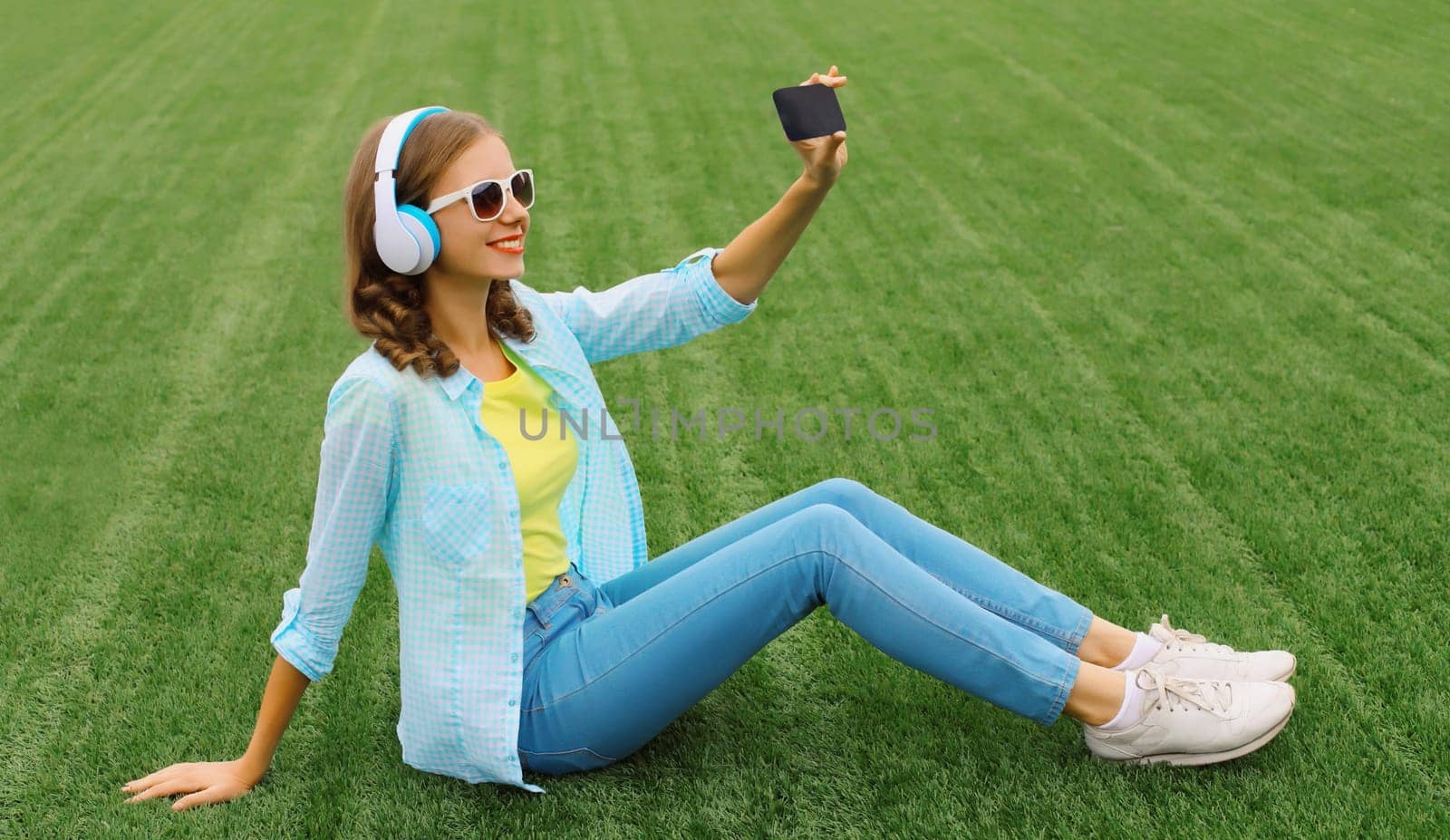 Happy young woman taking selfie with smartphone listening to music in headphones while lying on grass in summer park