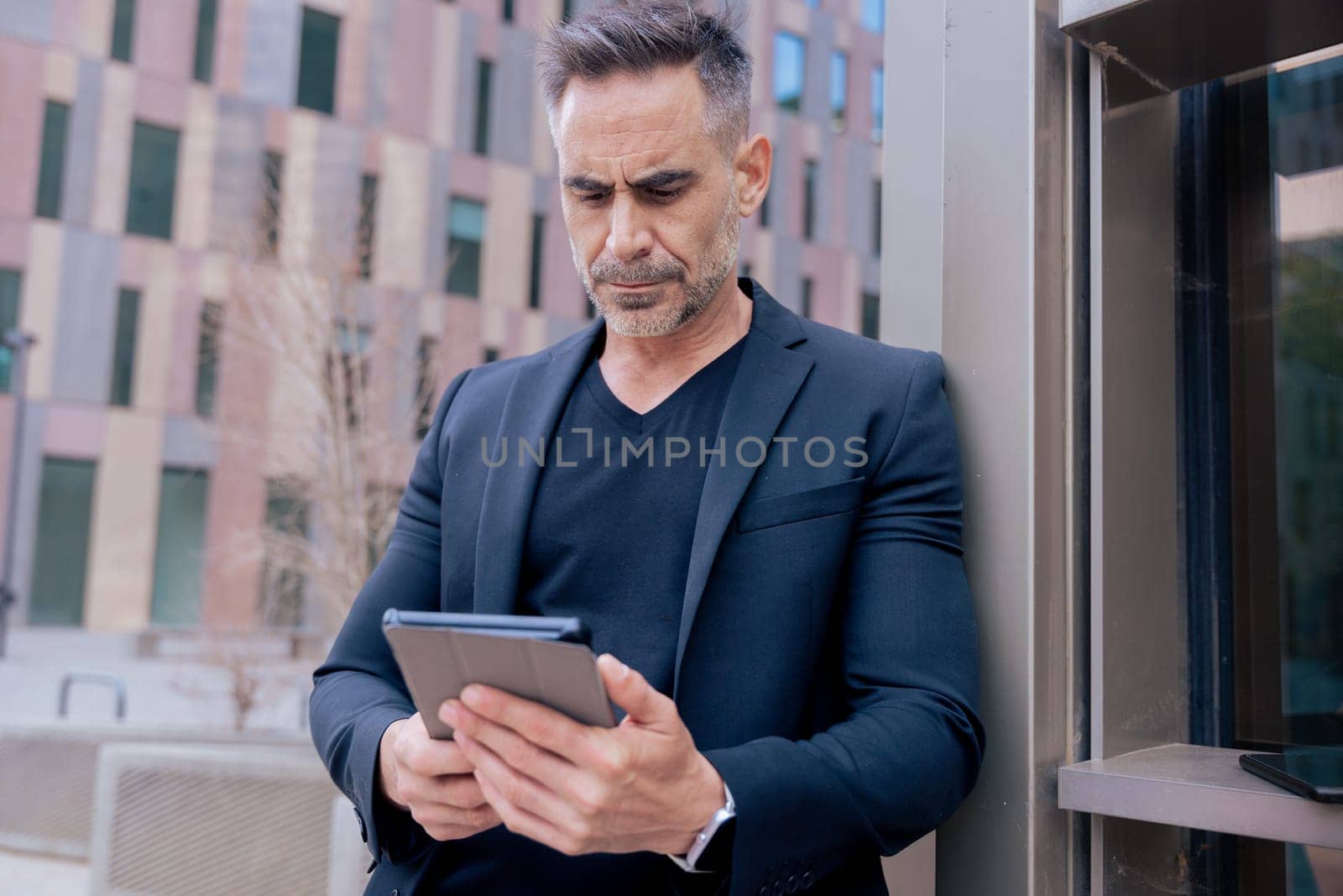 Confident mature businessman working on tablet outside a modern office building. Urban setting Ideal for business, technology, productivity, and professional lifestyle themes.