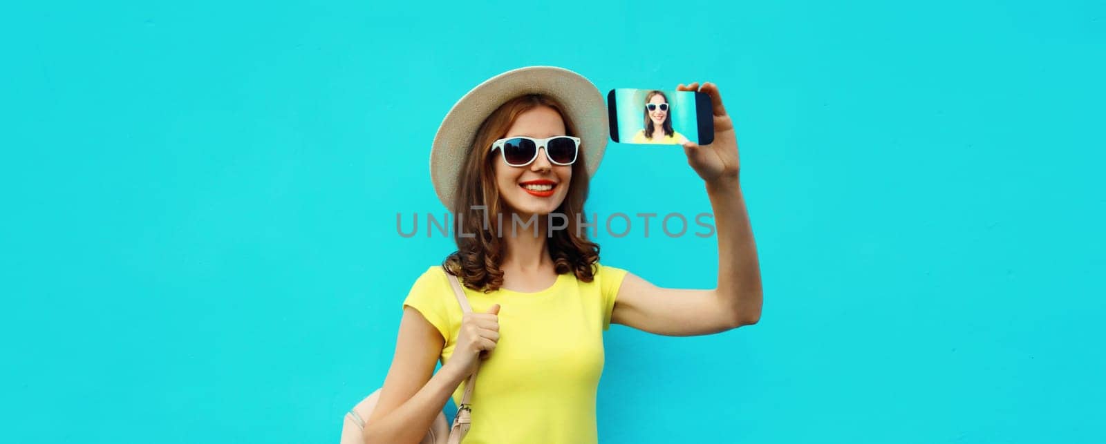 Portrait of happy smiling young woman taking selfie with smartphone wearing a summer straw hat with backpack on blue background