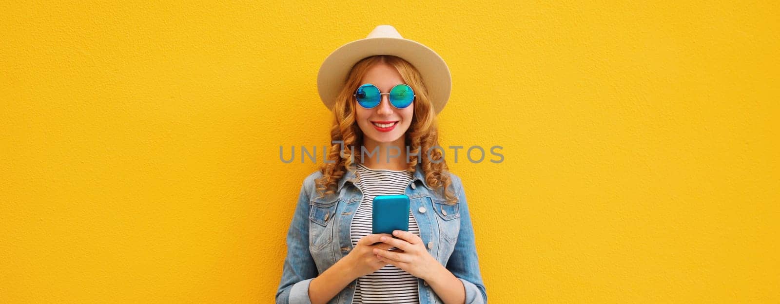 Summer portrait of happy traveler young woman with mobile phone looking at smartphone wearing a straw tourist hat, casual clothing on yellow studio background