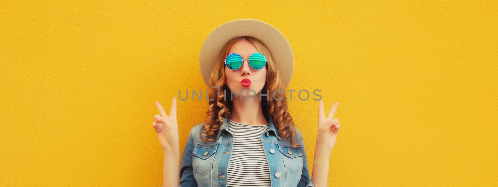 Portrait of beautiful stylish young woman blowing kiss in summer hat, glasses, denim jacket posing on yellow studio background