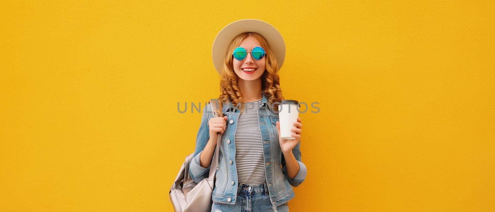 Beautiful stylish happy smiling young woman with cup of coffee wearing summer hat, backpack, jean jacket on bright wall yellow studio background
