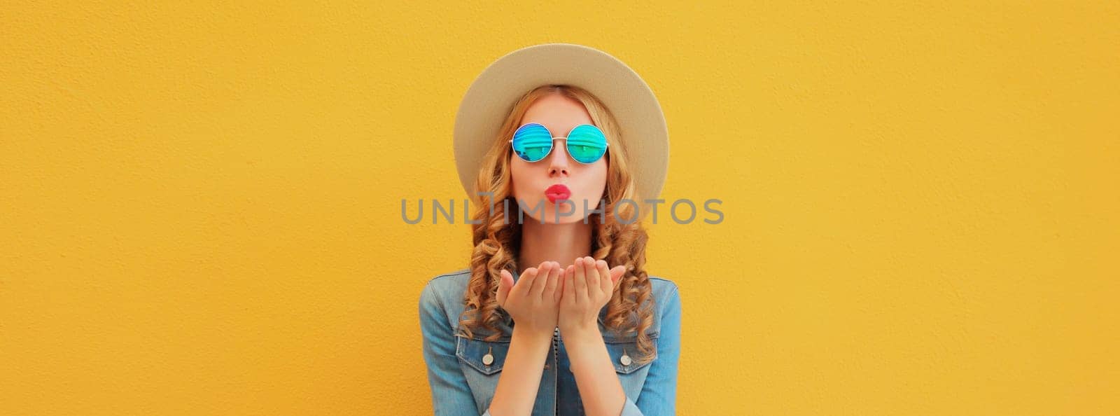 Portrait of beautiful stylish young woman blowing kiss in summer hat, glasses, denim jacket posing on yellow studio background