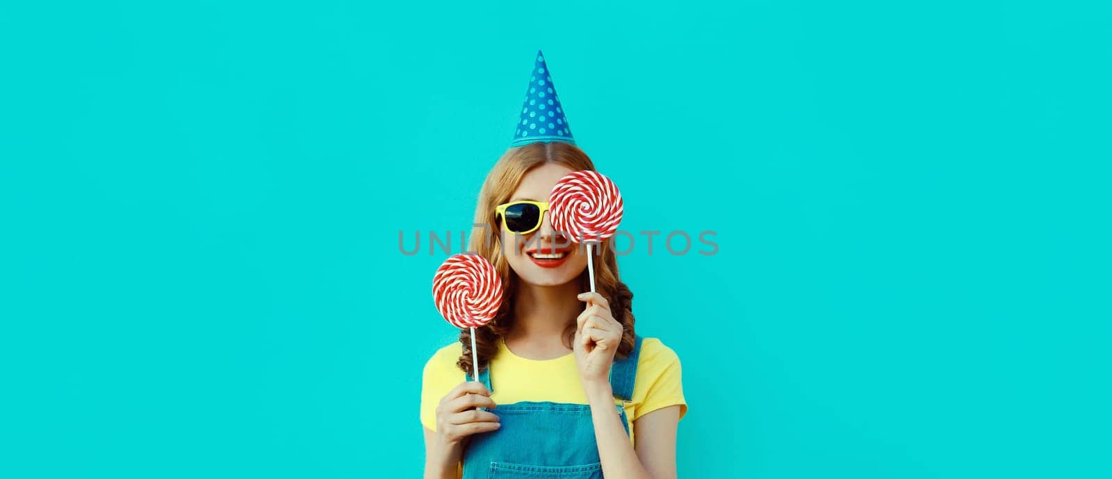 Holiday, celebration, event. Happy cheerful young woman laughs celebrating having fun at a party wearing festive birthday hat holding lollipop on blue background