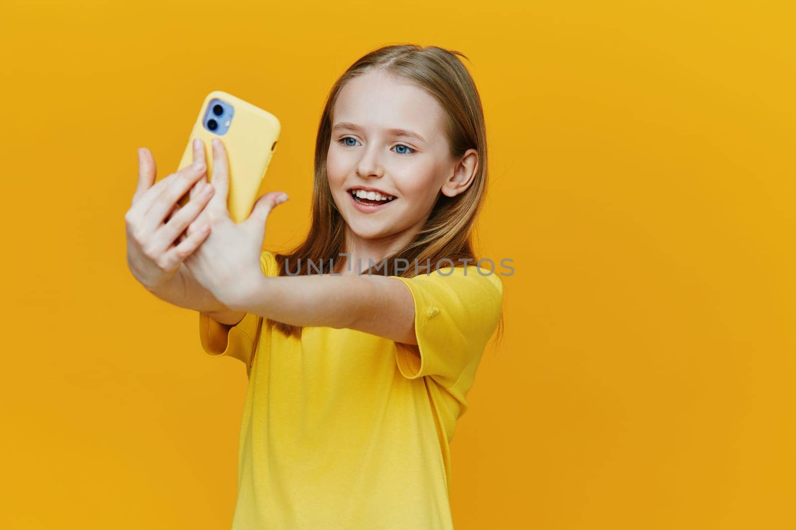 A cheerful young girl captures a selfie with a yellow smartphone, expressing happiness against a vivid yellow backdrop, perfect for summer themes and social media concepts