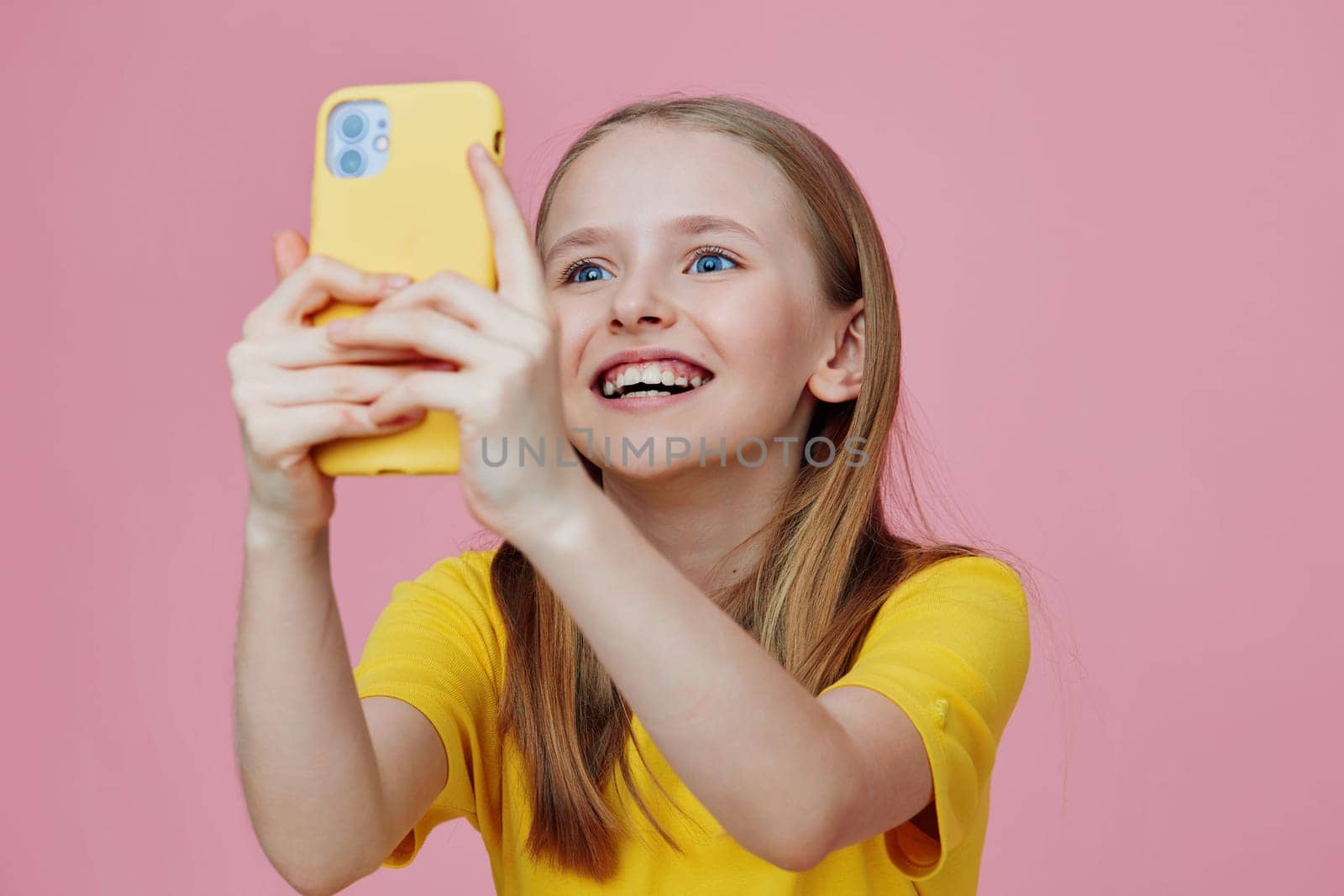 A joyful young girl with long hair smiles brightly while capturing a selfie on her yellow phone against a pink backdrop This image captures the essence of happiness, youth, and vibrant energy