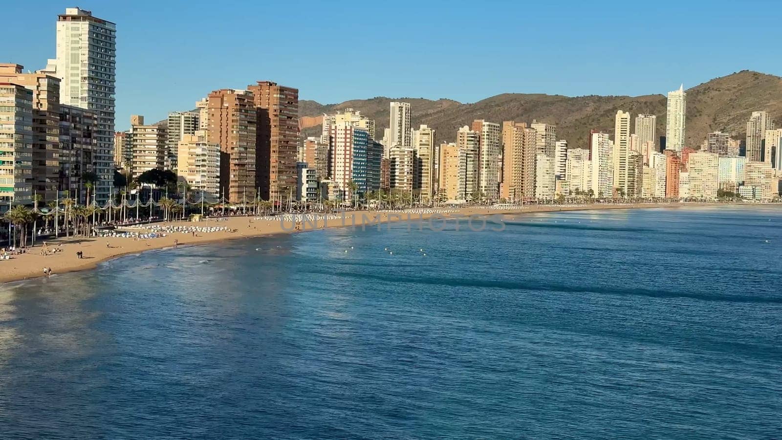 A breathtaking panoramic view of a coastal city with high rises, a lovely beach, and majestic distant mountains