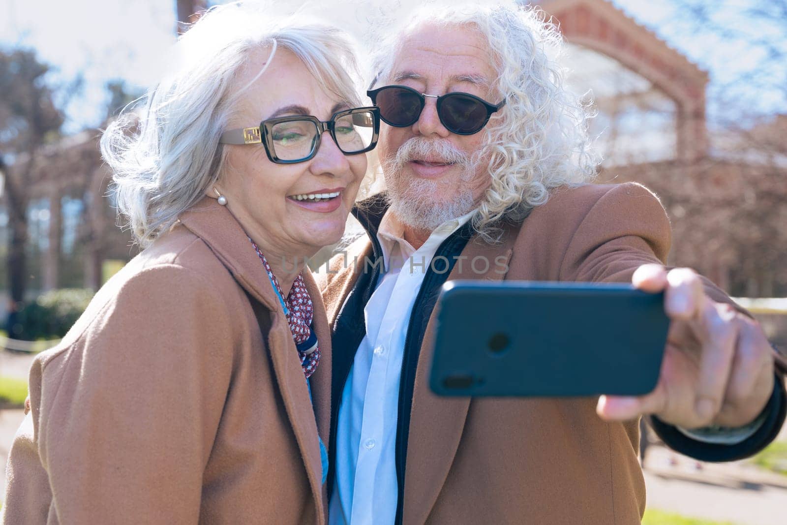 Happy Caucasian elderly couple sitting on a bench taking a selfie together outdoors. They wear brown jackets and glasses, enjoying a sunny day and capturing the moment with a smartphone.