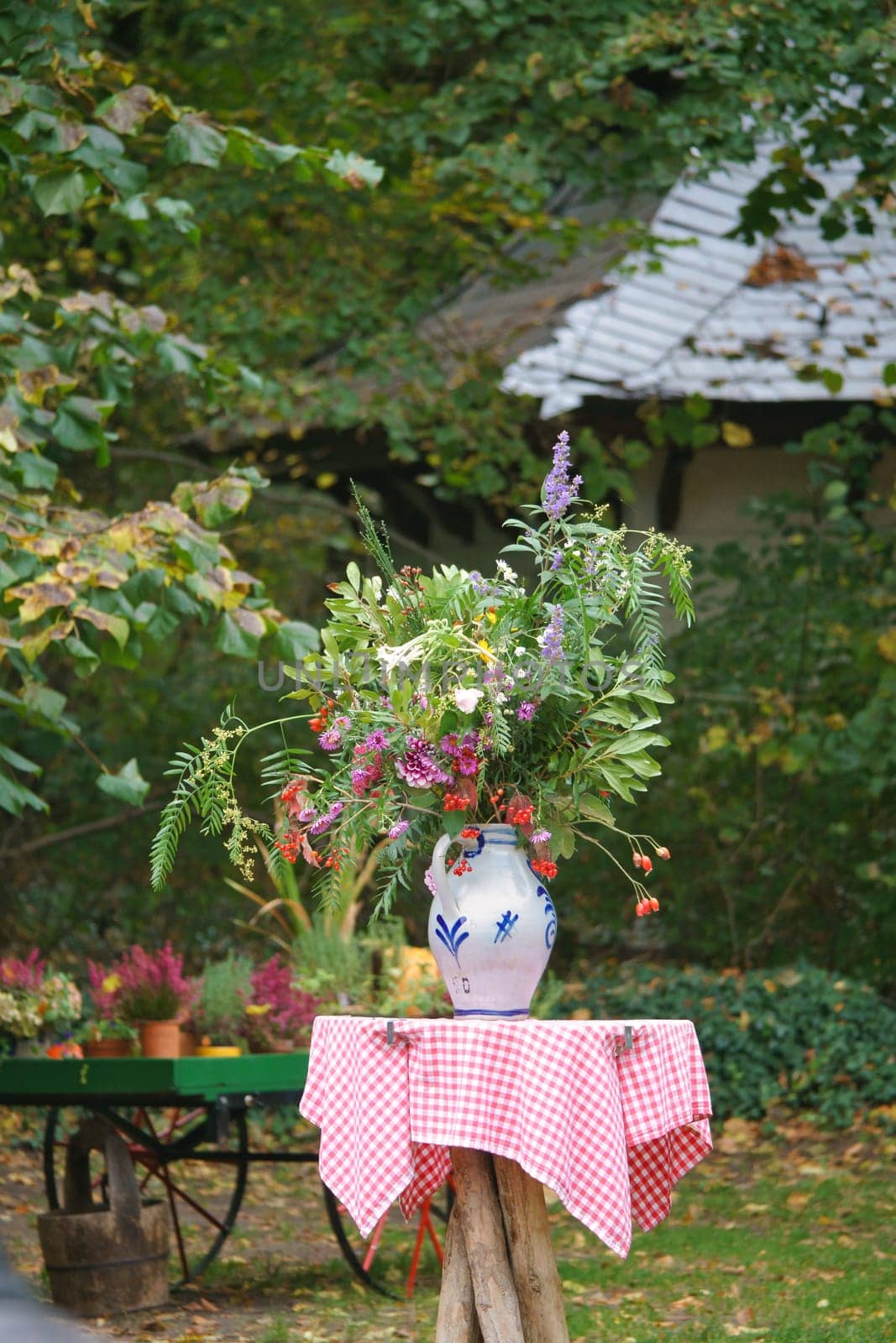 A bouquet of flowers in a vase on a table with a red and white checkered cloth, in the background a garden with trees and plants. Ideal for nature and garden lovers.
