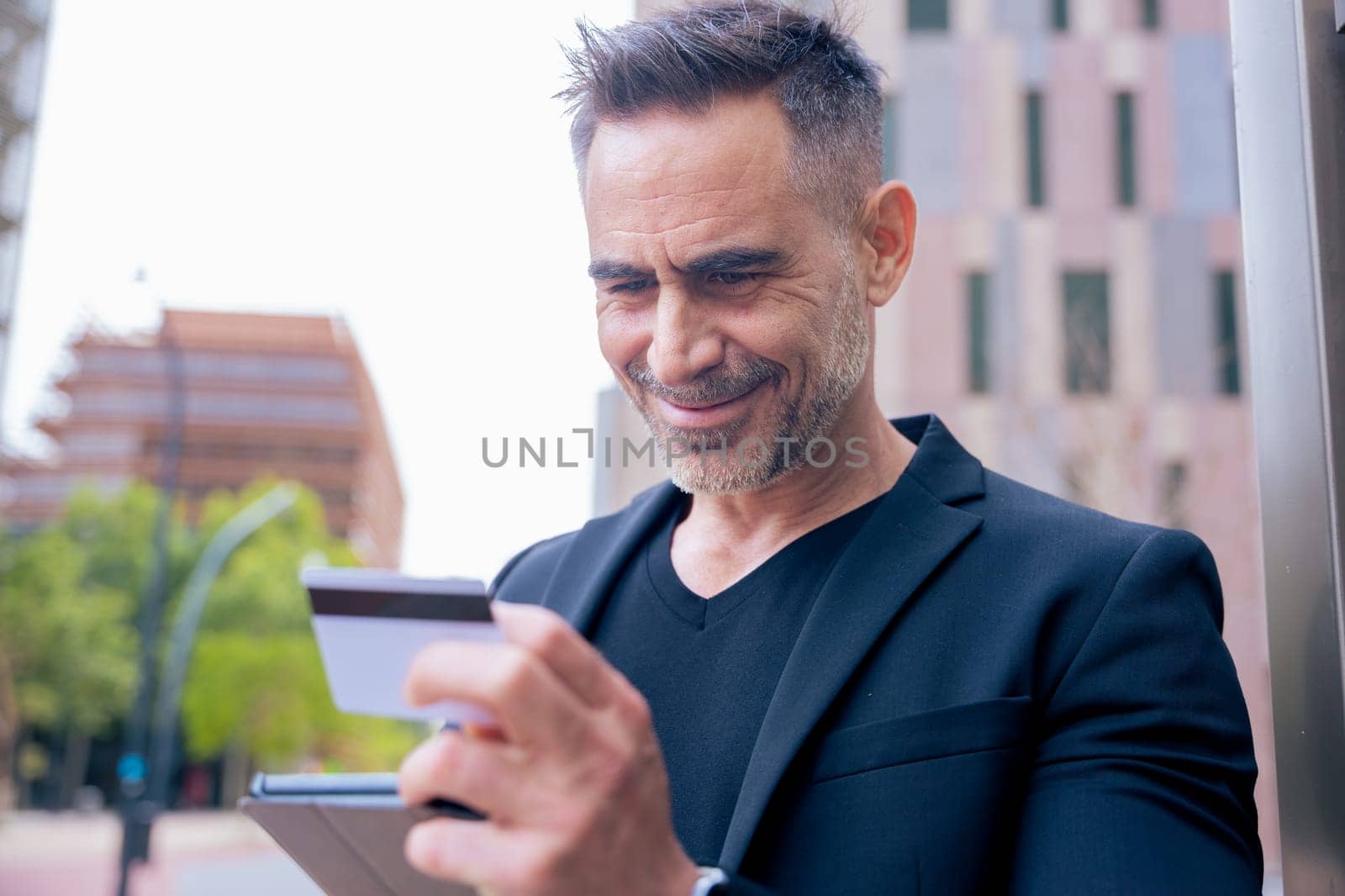 Confident caucasian man with headphones smiling while using tablet and holding a credit card outdoors. Concept of online shopping, digital payment, modern lifestyle, and financial technology.
