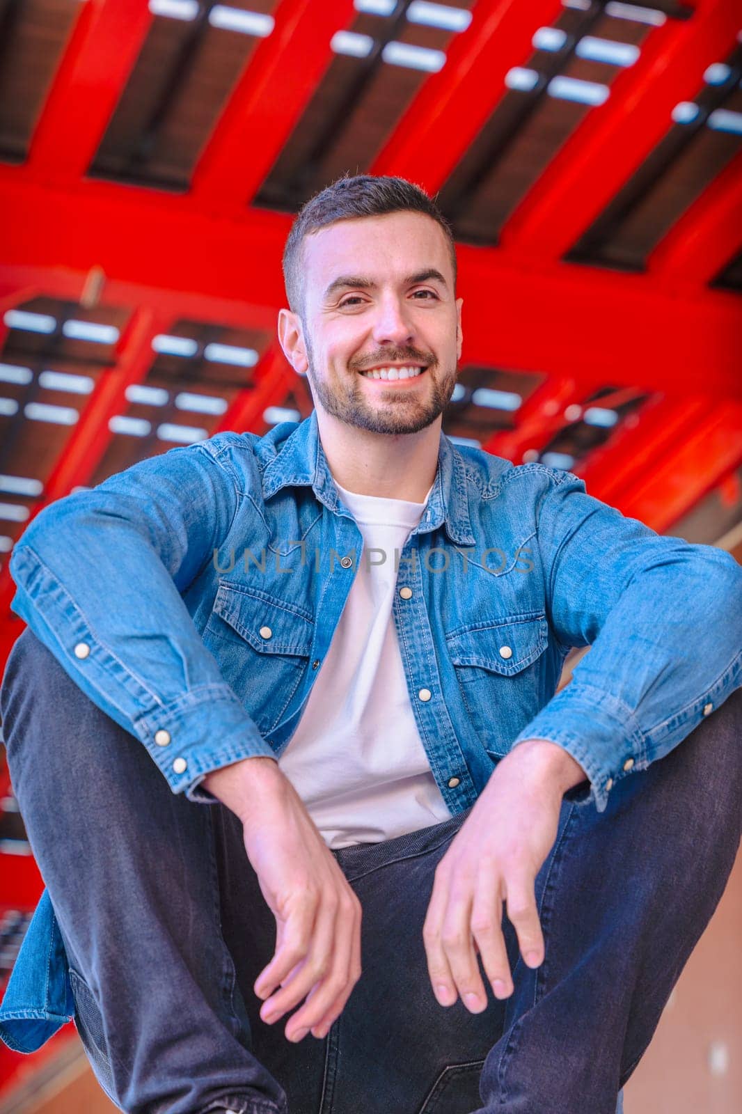 Vertical Portrait smiling young man sitting casually under a modern red urban structure. Relaxed pose, denim style, and confident expression in a bright. looking at camera