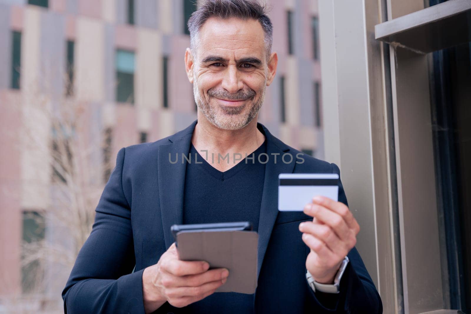 Confident caucasian man with headphones smiling while using tablet and holding a credit card outdoors. Concept of online shopping, digital payment, modern lifestyle, and financial technology.