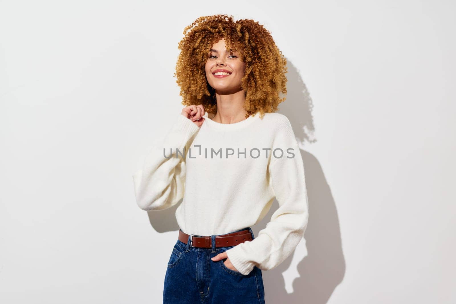 A cheerful young woman showcasing her natural curly hair while wearing a cozy white sweater and blue jeans She poses confidently with a smile, set against a clean, light background that highlights h