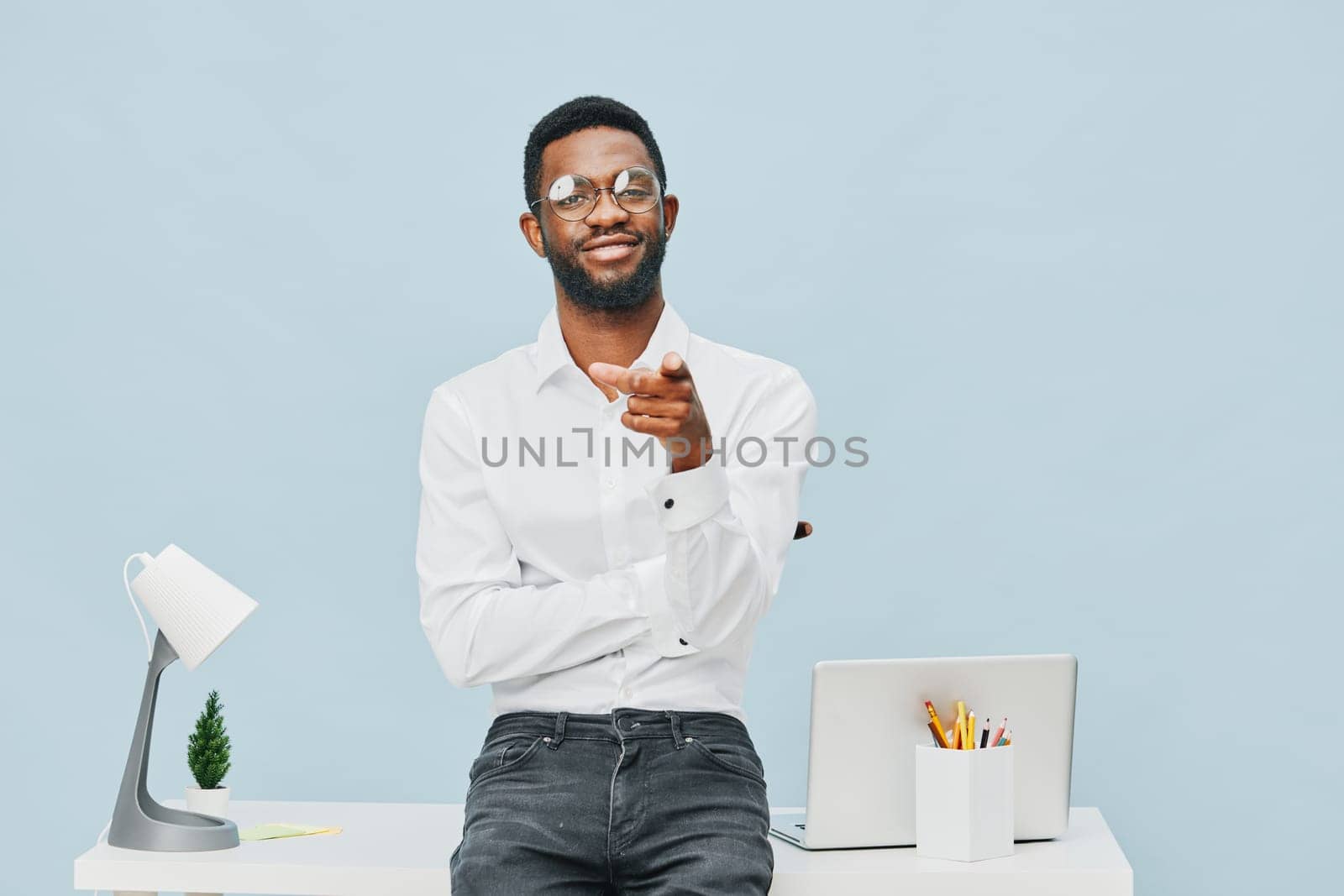 Cheerful confident man in casual office attire, wearing glasses and white shirt, standing at a desk with laptop, desk lamp, and potted plant in a bright, minimalistic workspace setting. Concept: professional success and positivity.