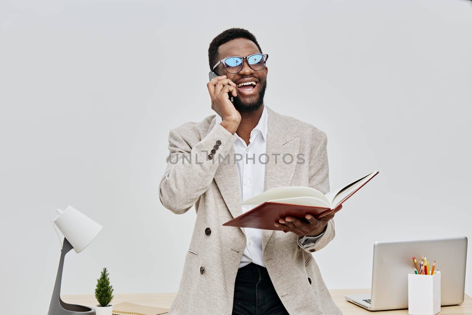 Smiling man with glasses using a smartphone and holding a notebook in a bright office setting with a minimalistic desk, laptop, and plant, displaying happiness and confidence, interior photography, professional business atmosphere, modern work environment. Concept: positive communication