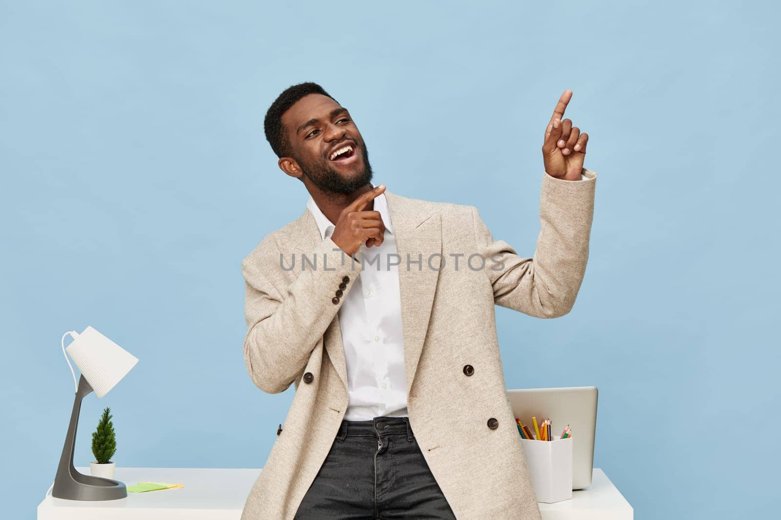 Happy energetic man in business casual clothes standing in bright contemporary office, gesturing with finger upward, cheerful mood, blue backdrop, workspace with computer, stationery, and plant, work life style concept.