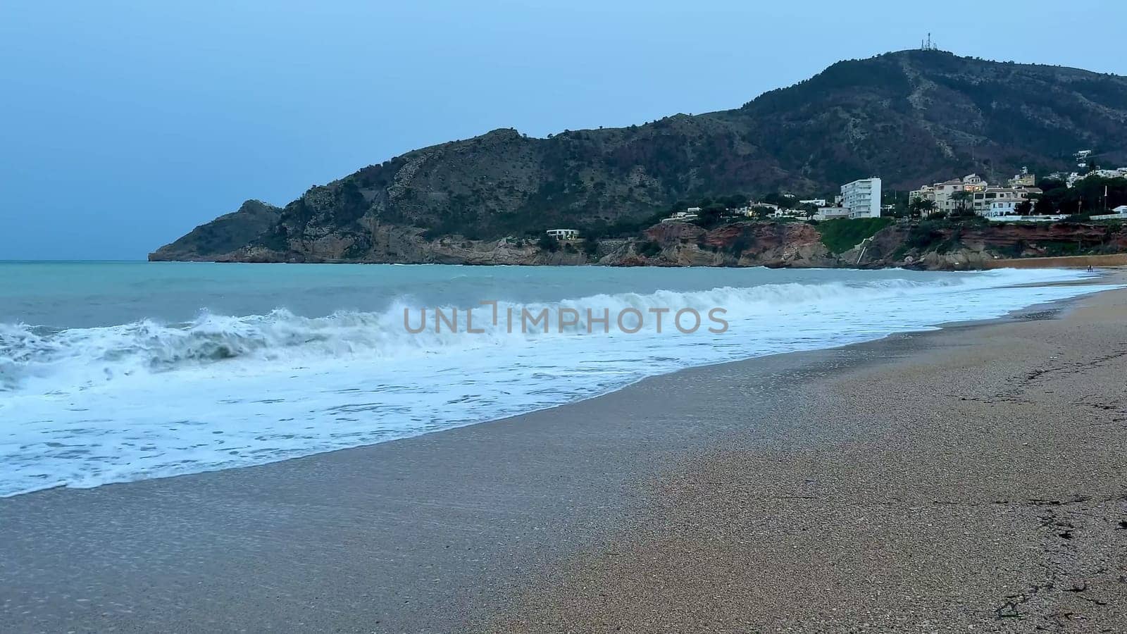 Calm and gentle waves are softly lapping on a sandy beach, framed by a stunning mountain backdrop at dusk