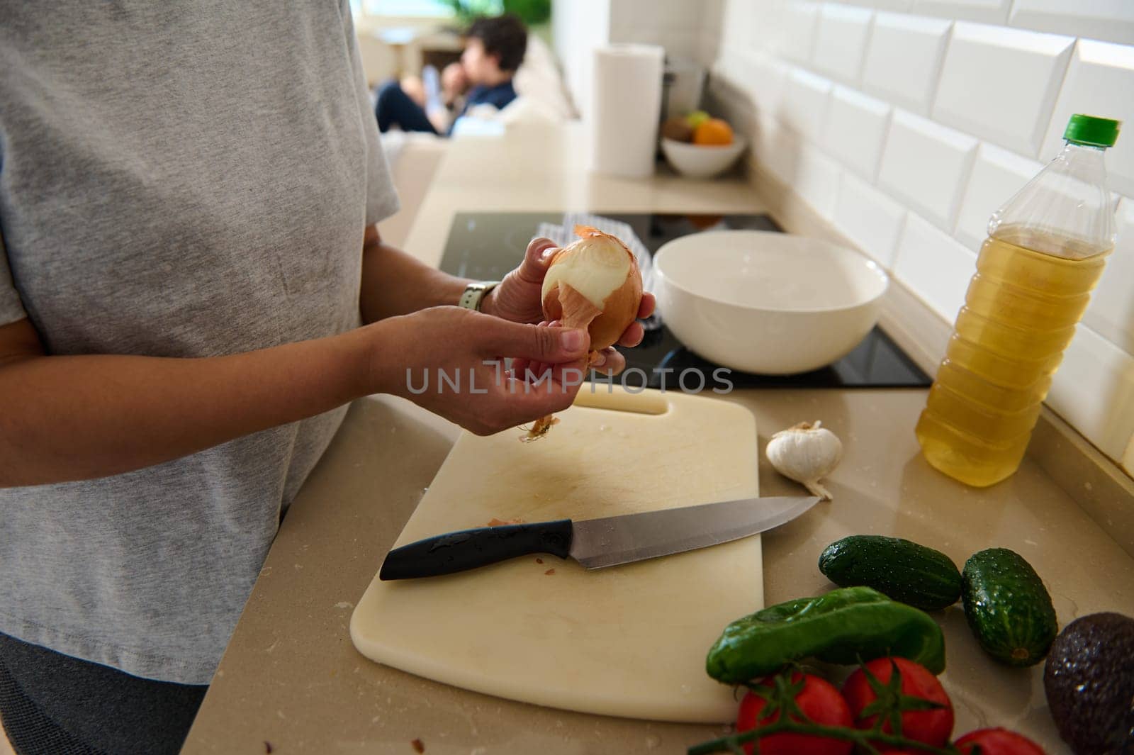 A person peels an onion in a contemporary kitchen with fresh ingredients laid out, including tomatoes and cucumbers. A detail view emphasizing culinary preparation and healthy eating.