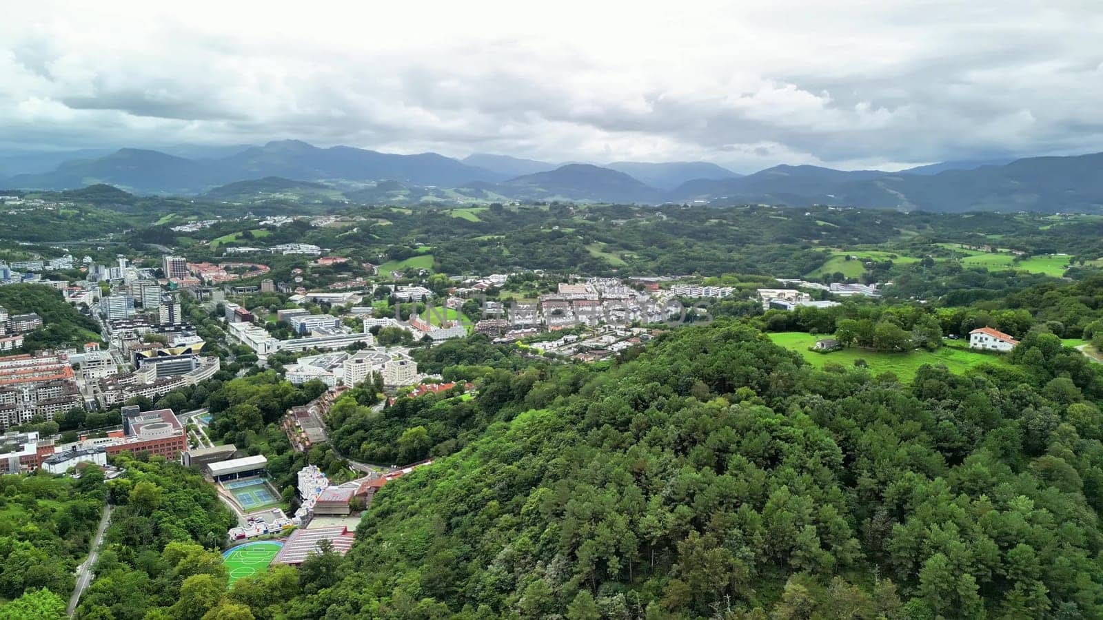 This aerial view highlights a coastal city framed by tall mountains and lush greenery surrounding it