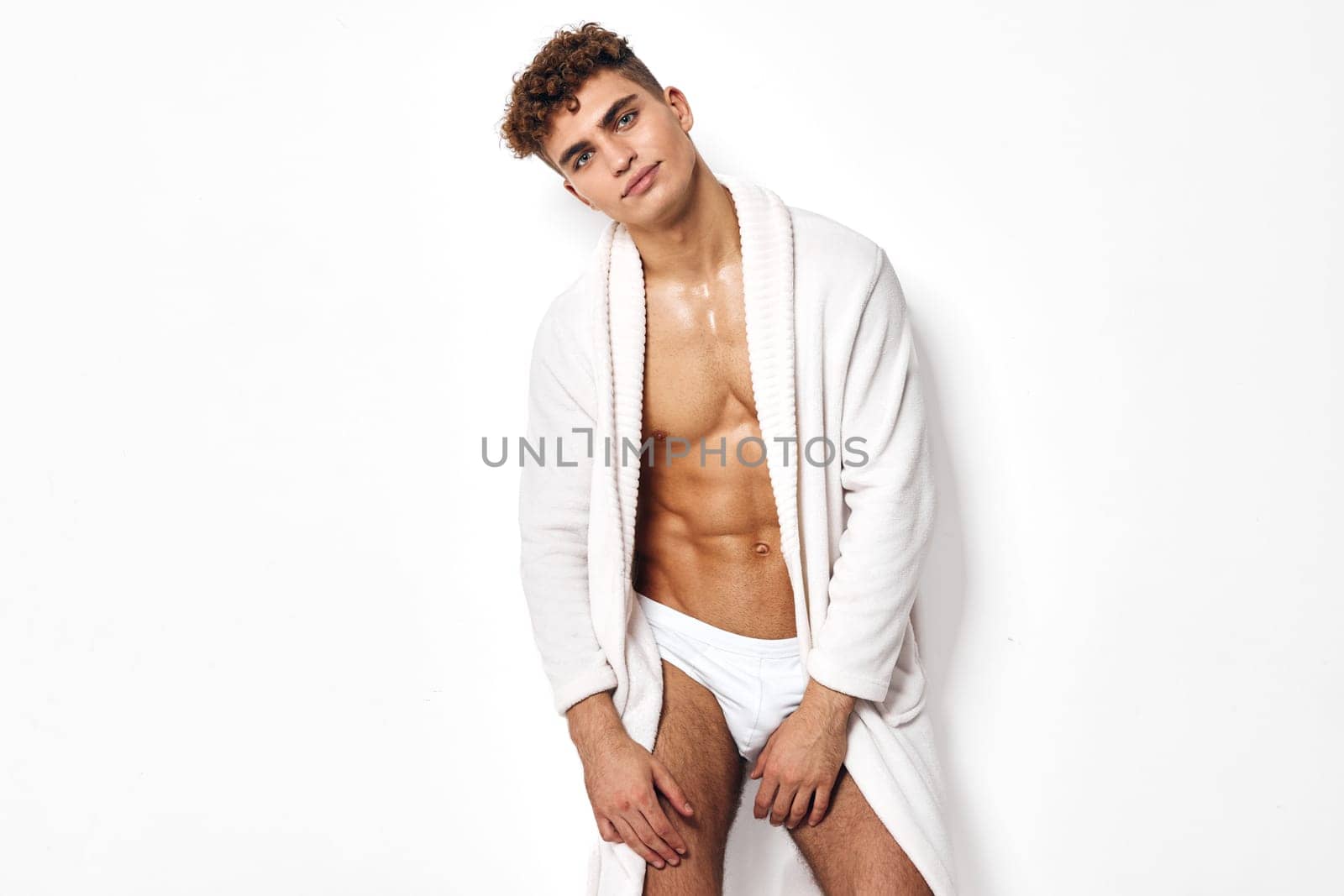 Young muscular man in a white bathrobe exuding confidence and relaxation. The pure white background emphasizes the theme of wellness and personal grooming.
