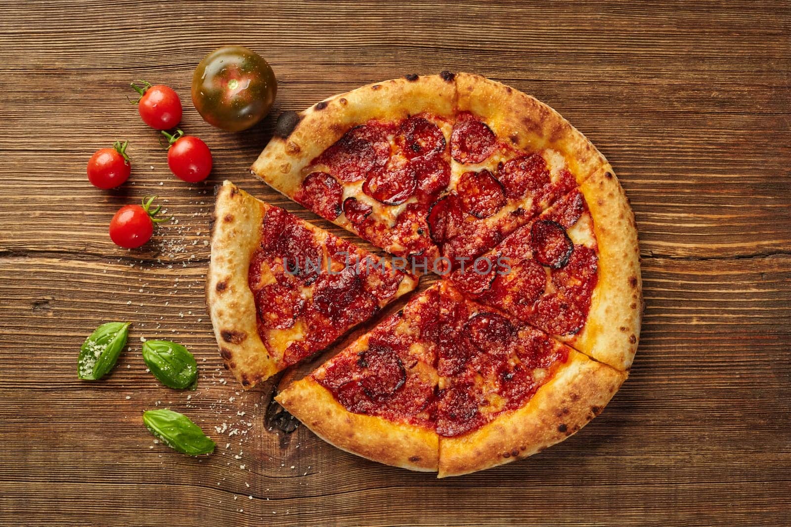 Sliced pepperoni pizza sits on a wooden table, surrounded by fresh tomatoes and basil leaves