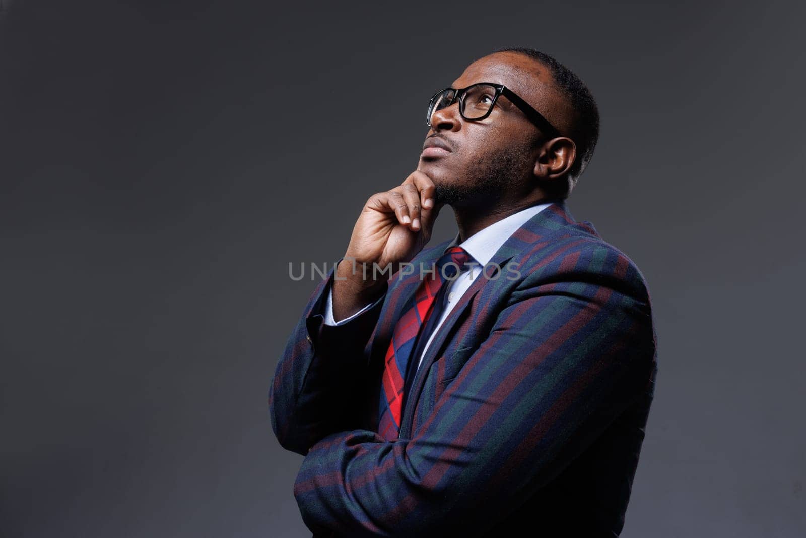 Thoughtful businessman in striped suit and glasses, looking up with hand on chin. Concept of decision-making, planning, strategy, leadership, and innovation. Dark studio background.