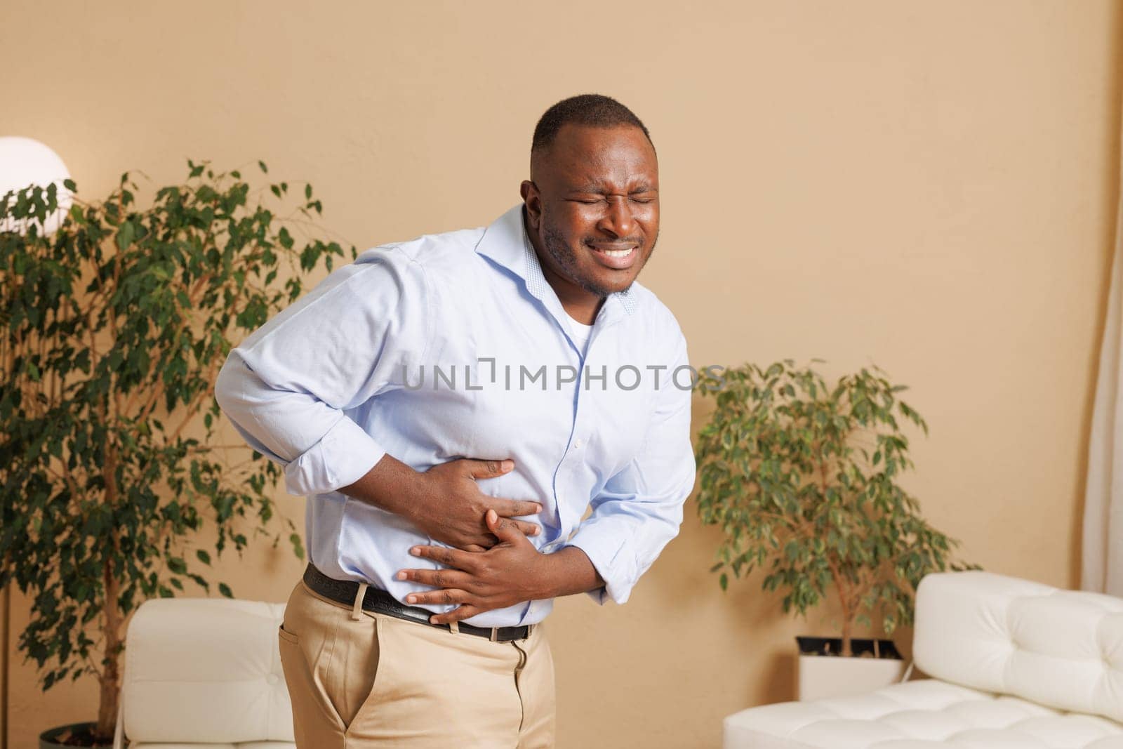 Man suffering from severe stomach pain at home. He clutches his abdomen with a pained expression, indicating digestive issues, cramps, or food poisoning. Health and medical concept.