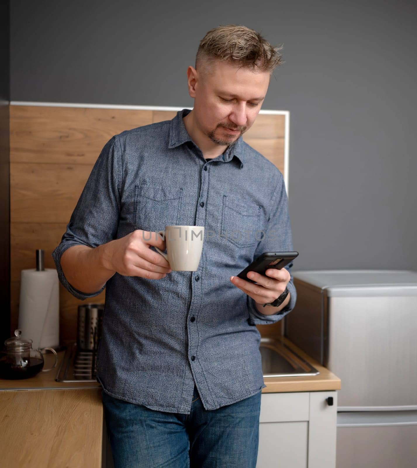 Handsome Man Drinks Coffee In The Kitchen And Watches The News On Phone