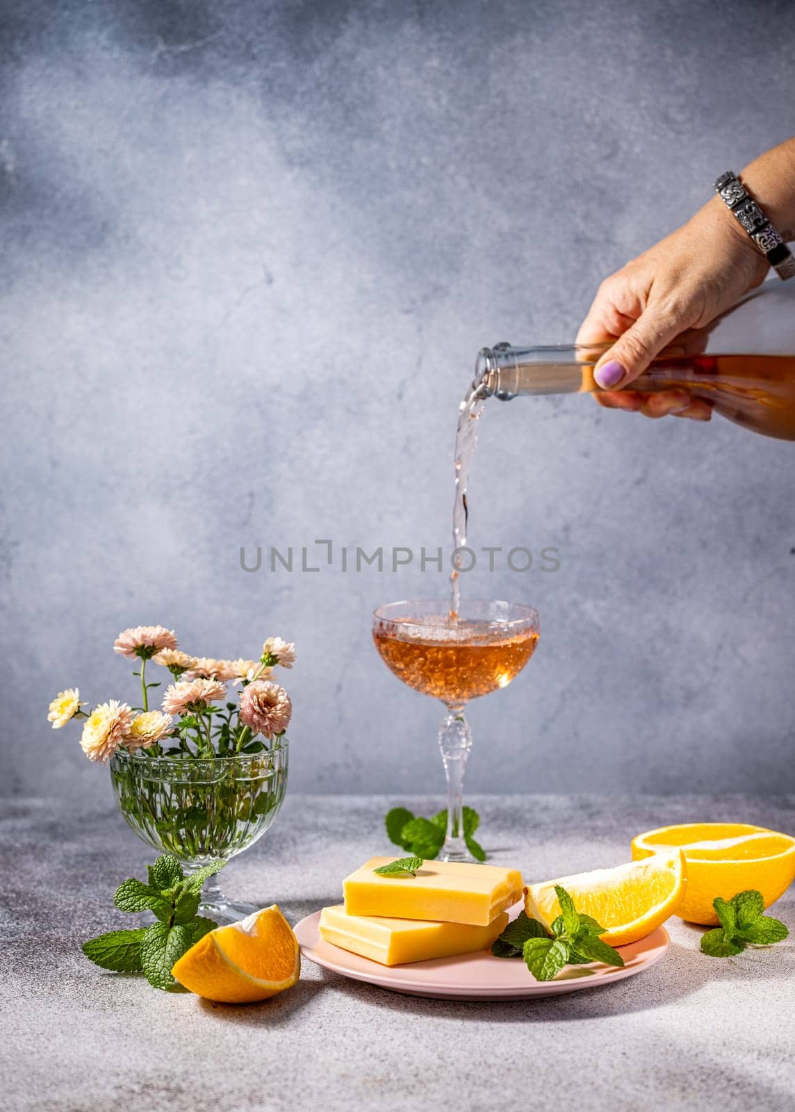 Rose wine being poured into a glass, accompanied by cheese, orange slices, and mint, creating a sophisticated and refreshing scene