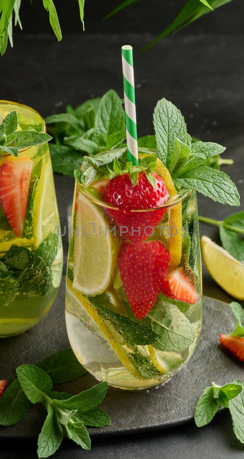 A refreshing drink in a glass with lemon slices, strawberries, fresh mint leaves, and sparkling water, served with lime and mint on a black plate