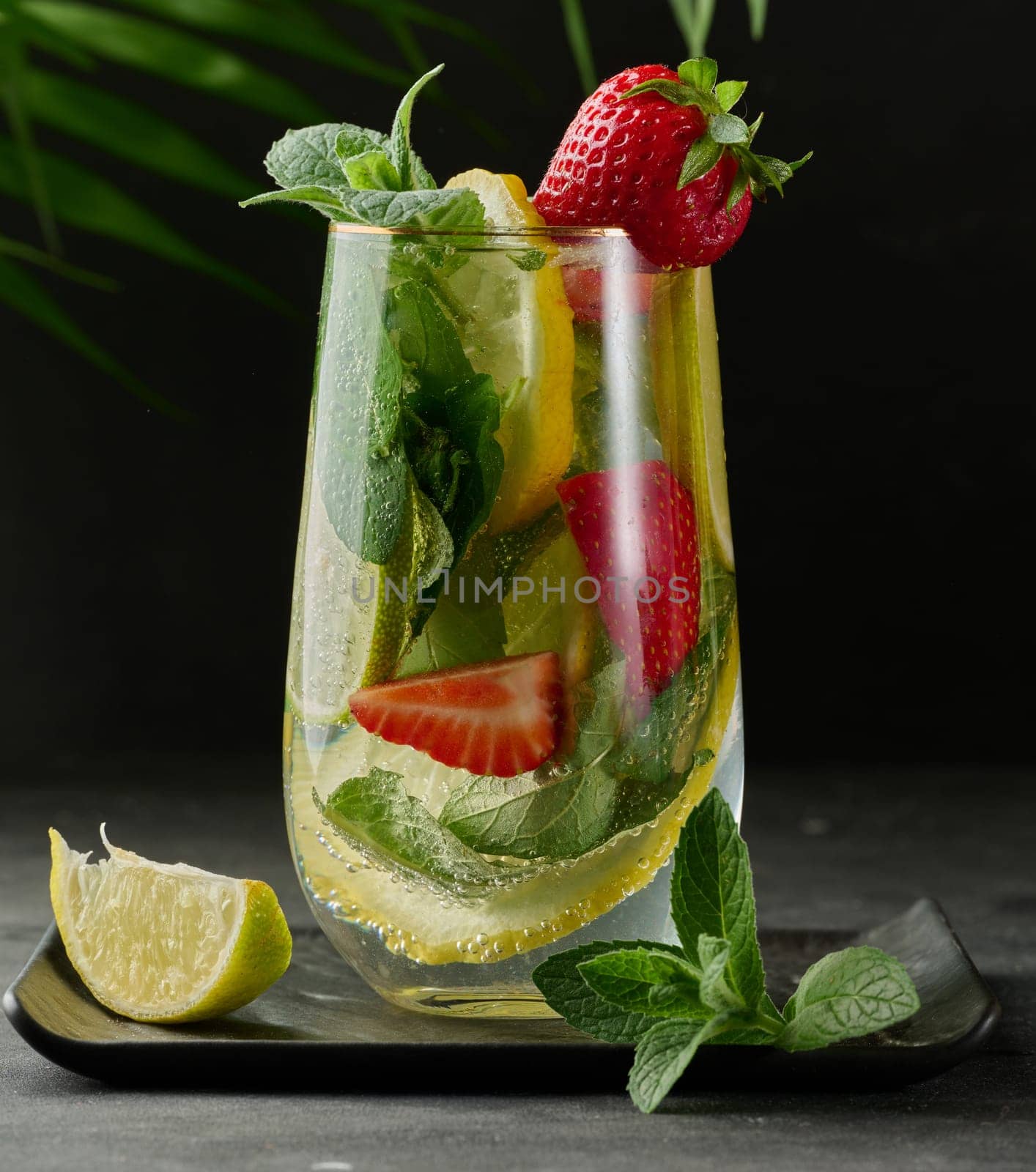 A refreshing drink in a glass with lemon slices, strawberries, fresh mint leaves, and sparkling water, served with lime and mint on a black plate.