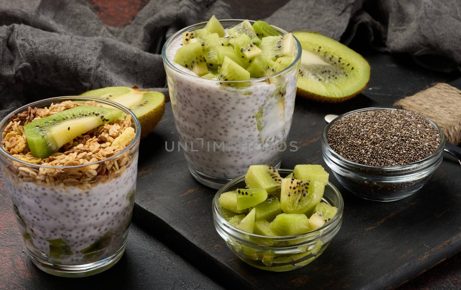 Two glasses of chia seed pudding, one topped with diced kiwi and the other with granola and banana chips, are on a dark surface with ingredients