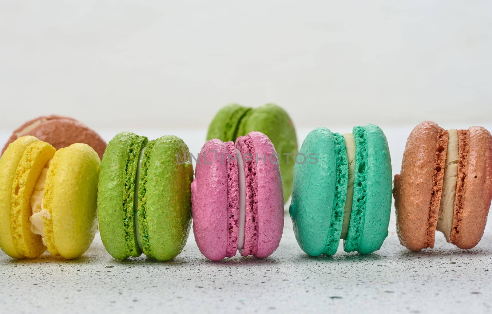 An assortment of colorful macarons on a white surface with dark speckles. The macarons have a smooth, delicate shell and a creamy filling