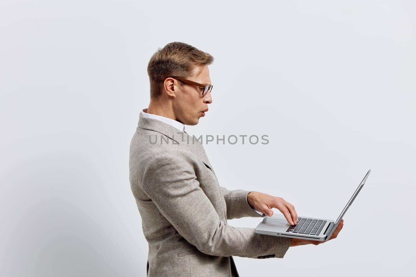 surprised man holding laptop wearing glasses and gray blazer on plain white background. young caucasian man with casual hairstyle using laptop for work in digital nomad lifestyle concept. innovation, remote work, connectivity.