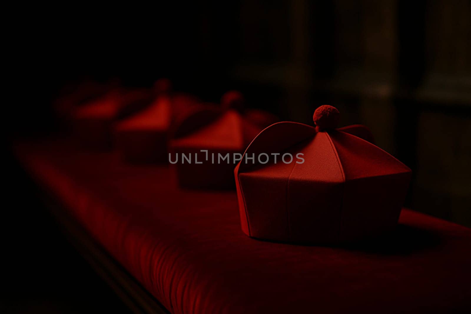 Red cardinal birettas arranged on a velvet bench inside a shadowed church interior, evoking solemnity and the atmosphere of a papal conclave