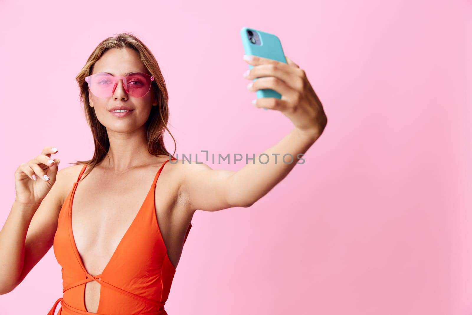 Vibrant woman in orange swimsuit capturing a selfie on her cellphone against a soft pink backdrop