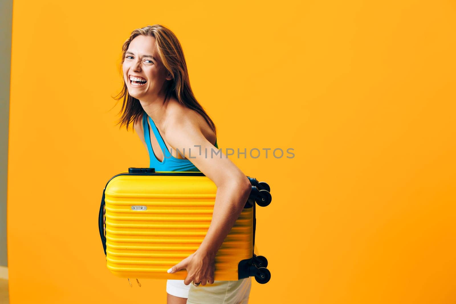 Cheerful woman holding a vibrant yellow suitcase standing against a bold orange wall with a joyful expression