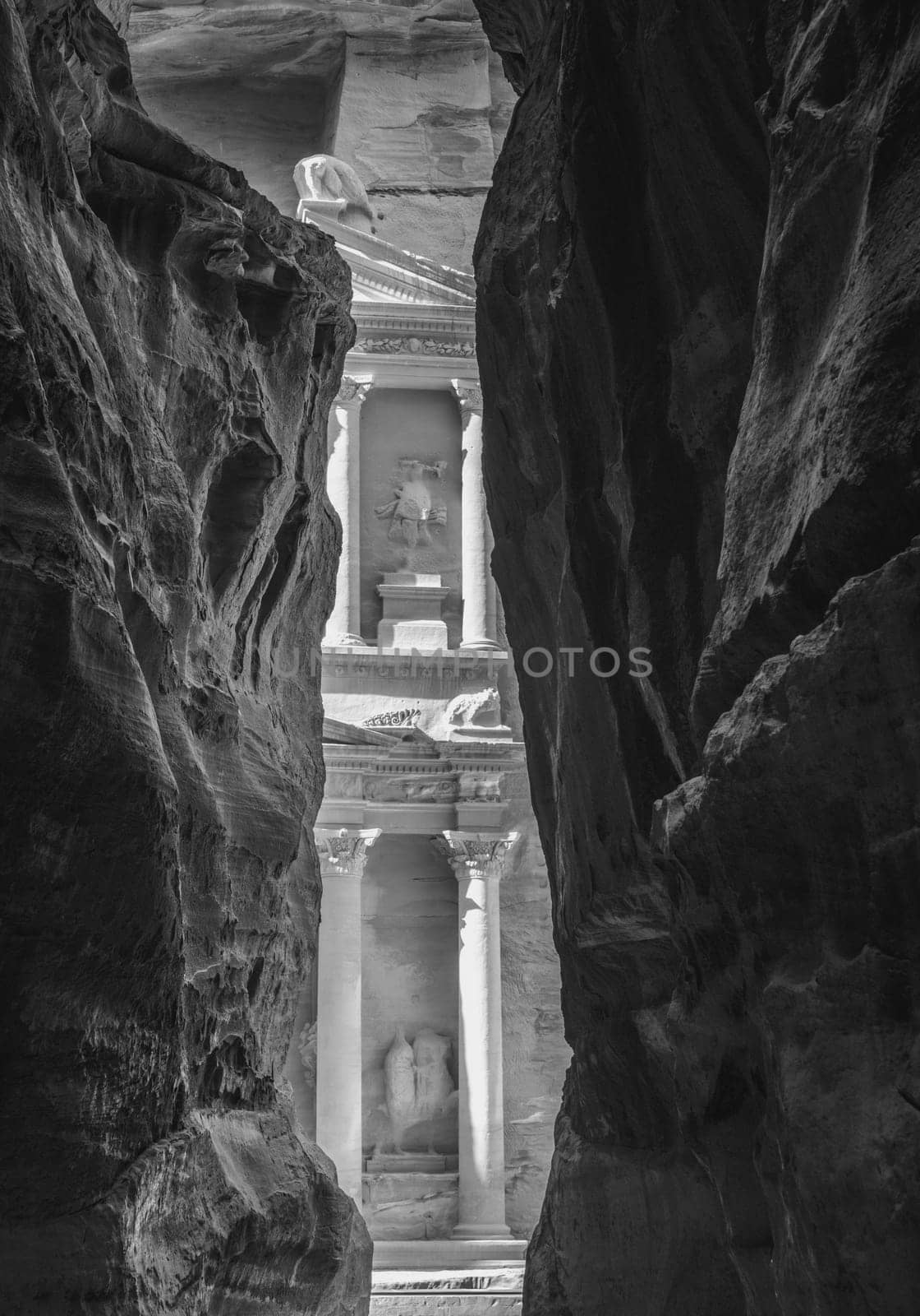 Black and white view of carved facade of the Treasury building or Al-Khazneh in the city of Petra in Jordan seen through narrow Siq gorge